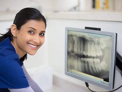 A dental hygienist is smiling at the camera while standing in front of a computer monitor displaying an X-ray.