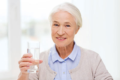 The image shows a woman holding a glass of water to her lips, smiling at the camera.