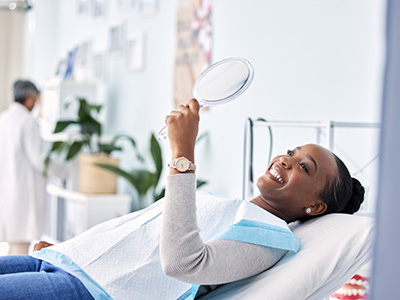 A woman sitting in a hospital bed, smiling and holding up a mirror to her face.