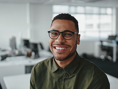 A man wearing glasses, smiling, and sitting in an office environment.