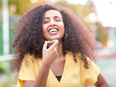 A woman with curly hair and a smile, wearing a yellow top, stands outdoors.