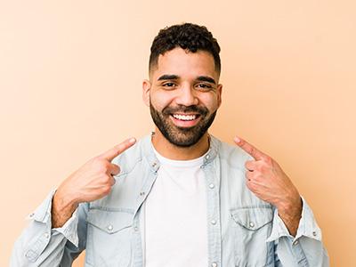 A young man with a beard and mustache, smiling broadly, pointing to his chest with one hand, against a light yellow background.