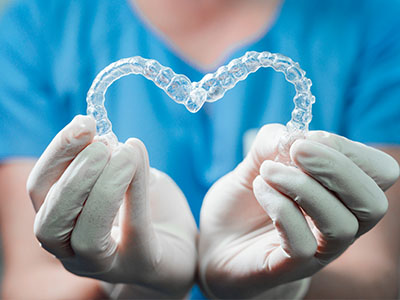 A dental professional holds a transparent, heart-shaped dental appliance in their gloved hands.