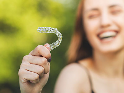 The image shows a young woman holding up a clear plastic toothbrush with her left hand, smiling and looking directly at the camera.