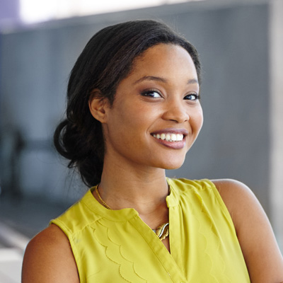 A smiling woman with dark hair, wearing a yellow top and posing against a background that appears to be an urban outdoor setting.