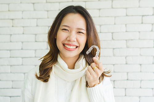 The image features a smiling woman holding up a toothbrush in front of her face. She is indoors with a brick wall behind her and appears to be standing against it.